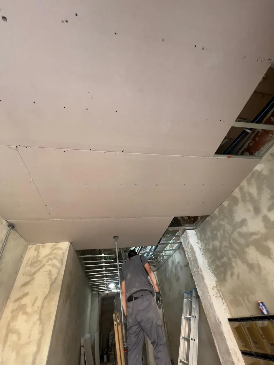 Un trabajador instala paneles de yeso en el techo de un pasillo interior estrecho, con una escalera cerca.