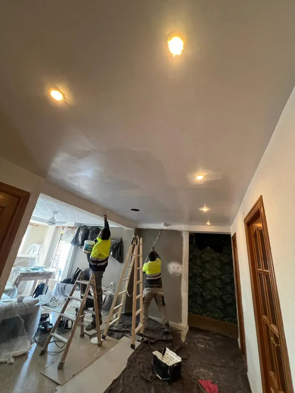 Dos trabajadores con chalecos amarillos subidos a escaleras pintan una habitación con luces empotradas en el techo.