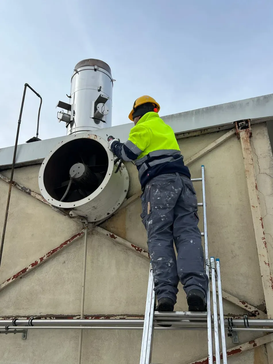 Un trabajador con chaqueta amarilla y casco repara una rejilla de ventilación industrial en la azotea mientras está subido a una escalera.