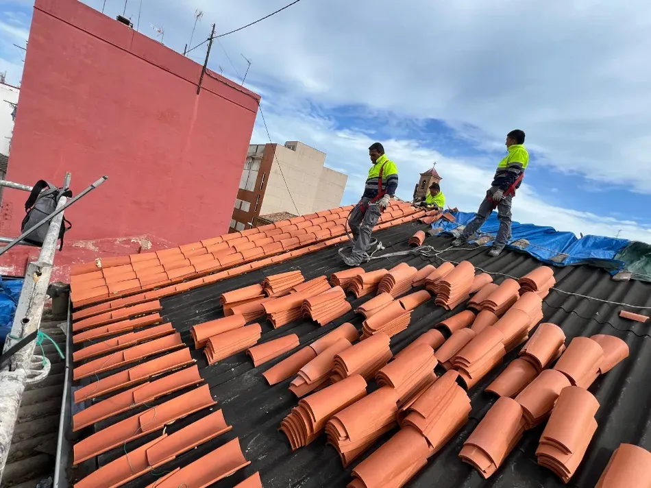 Obreros con chalecos reflectantes trabajando en un tejado de tejas durante las reparaciones, con un edificio rojo a su lado.