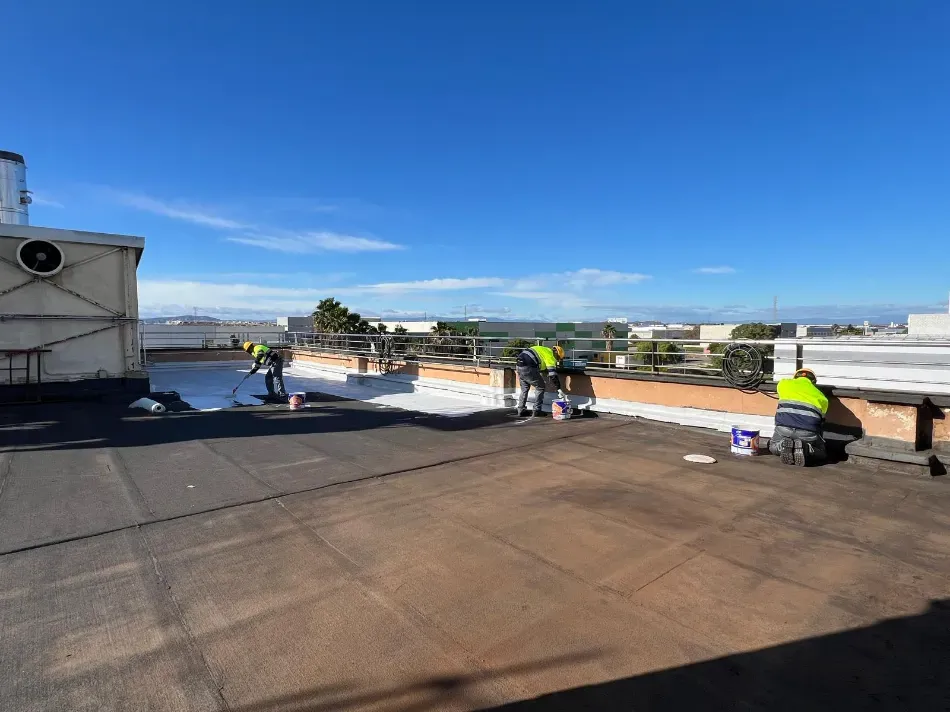 Trabajadores reparando una azotea plana bajo un cielo azul despejado, con herramientas y materiales esparcidos.