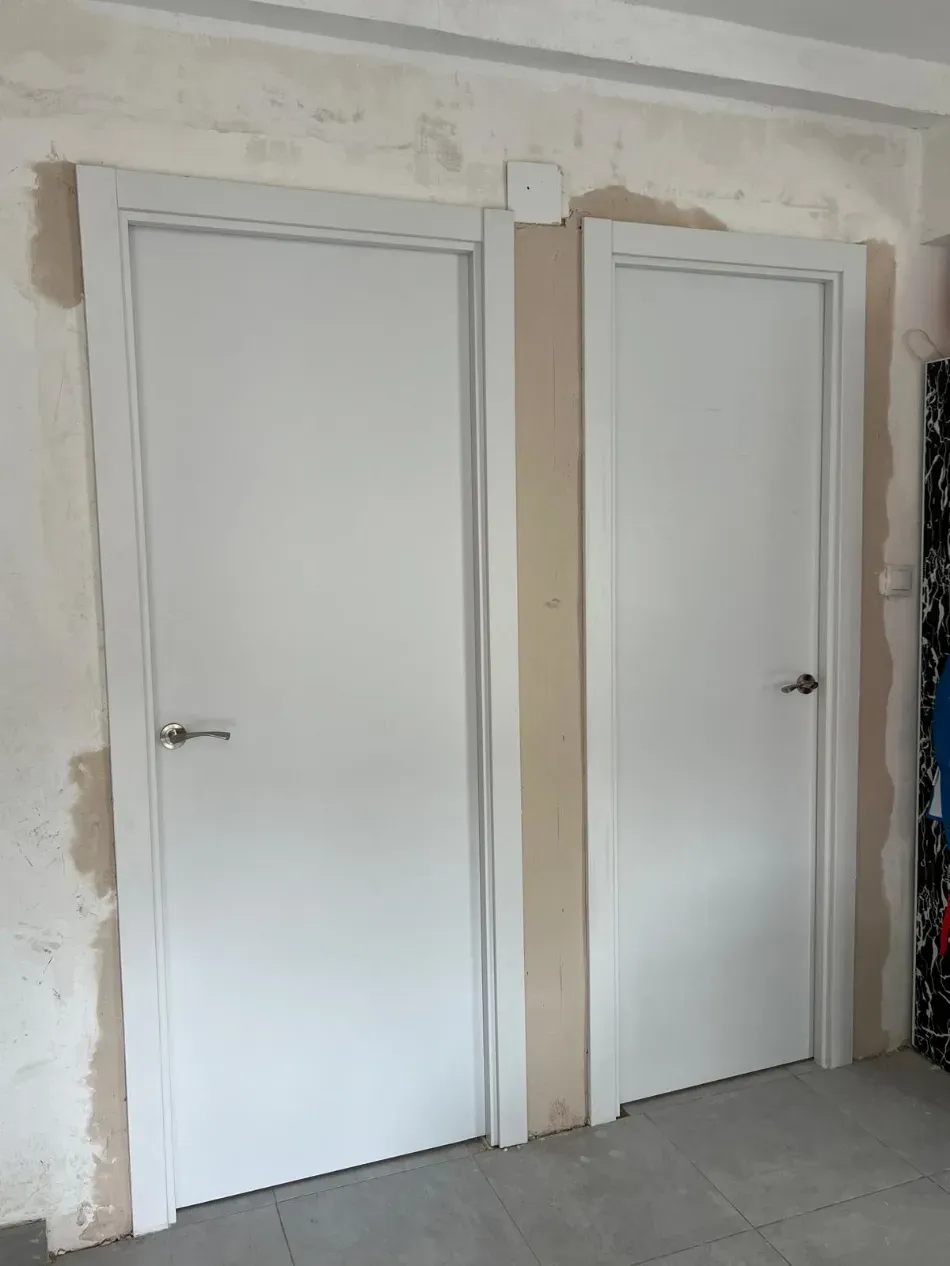 Dos puertas interiores blancas en una habitación con paredes sin terminar, con manchas y suelo gris.