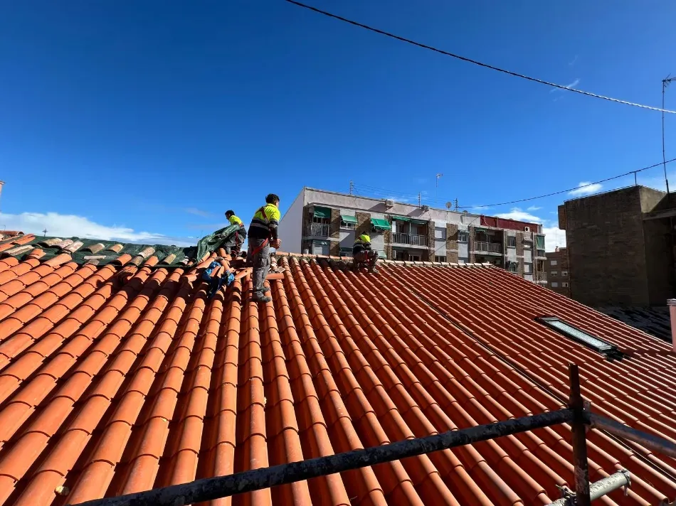 Trabajadores instalando o reparando tejas en un tejado de tejas rojas bajo un cielo azul despejado.