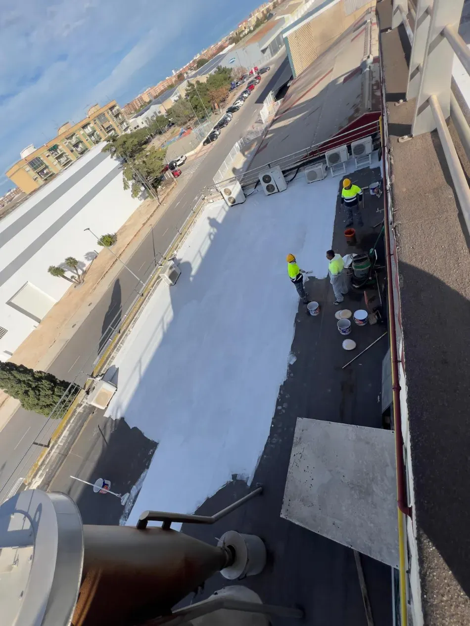Trabajadores con chalecos reflectantes reparan en una azotea una superficie negra junto a una rejilla de ventilación y reabastecen los suministros.