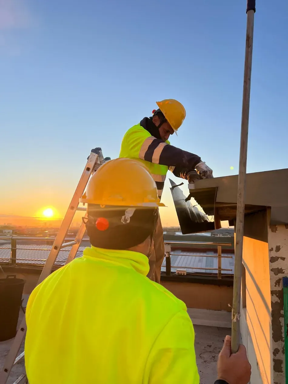 Dos trabajadores con cascos y chaquetas fluorescentes en una azotea al atardecer, uno pasándole una herramienta al otro.