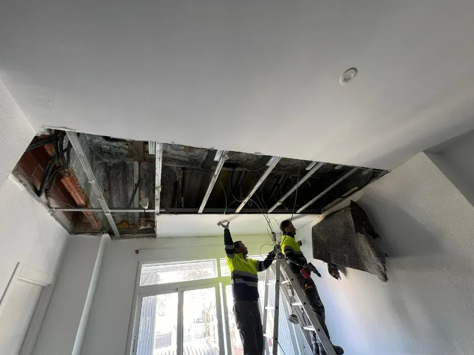 Dos trabajadores inspeccionan un techo dañado con vigas y cableado a la vista en una habitación luminosa.