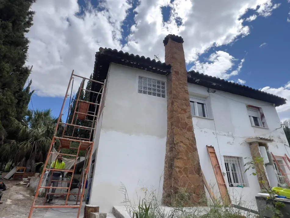 Exterior de una casa blanca con una alta chimenea de ladrillo y andamios a la izquierda bajo un cielo azul nublado.