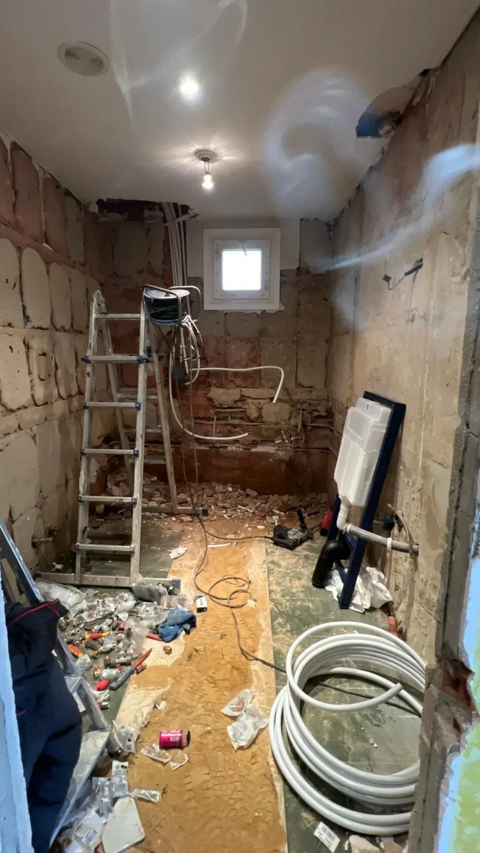 Habitación estrecha en obras con escaleras, herramientas, paredes a la vista, cableado y manguera enrollada en el suelo.