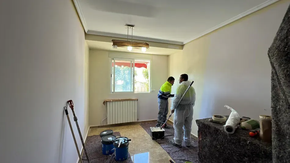 Dos trabajadores pintan una pequeña habitación con paredes blancas y una ventana; las herramientas de reforma están esparcidas por el suelo.