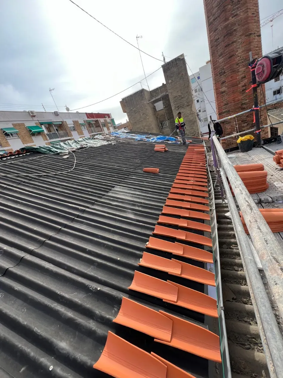 Hilera de tejas onduladas negras con cumbreras naranjas en una obra en construcción; un trabajador se ve a lo lejos.