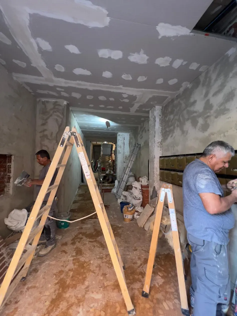 Obras de renovación en curso dentro de una habitación estrecha, con trabajadores, escaleras y paredes y techo de pladur sin terminar.