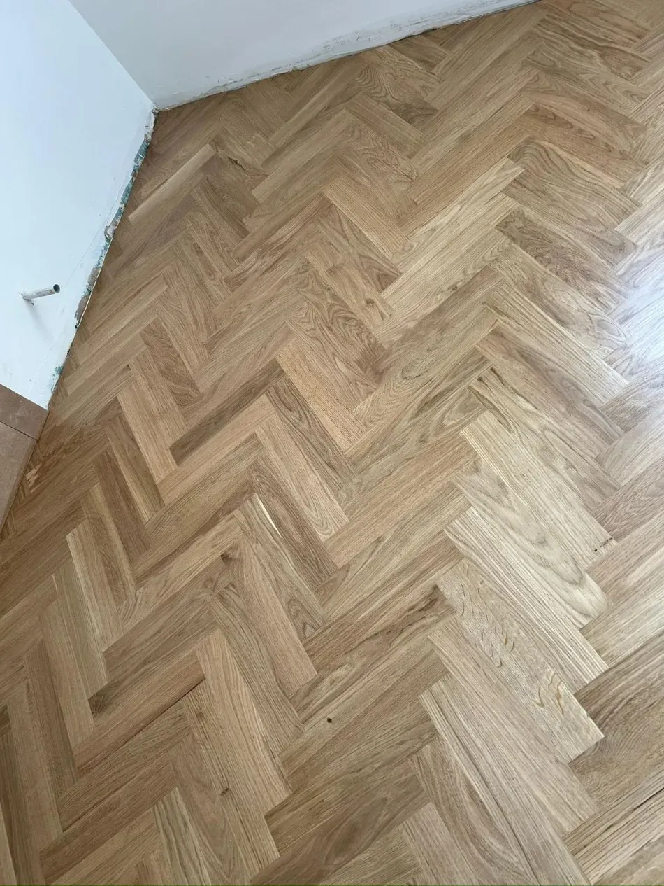 Habitación vacía con suelo de madera clara en espiga y paredes blancas.