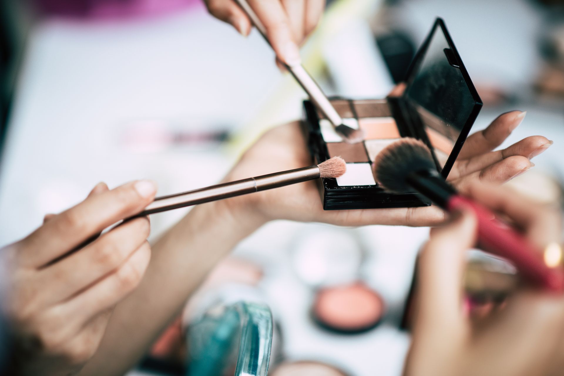 Des mains appliquant du fard à paupières avec des pinceaux de maquillage, à côté d’une palette de fards à paupières ouverte.