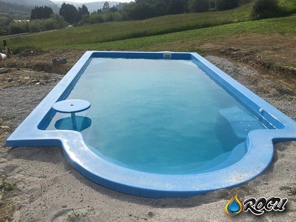 Piscina rectangular de color azul, con extremo curvo, pequeña mesa en la esquina, situada en una zona exterior con césped.