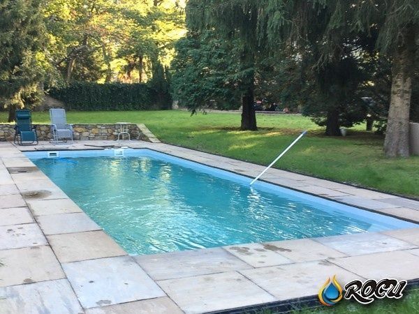 Una piscina rectangular rodeada de baldosas de piedra y césped verde, con árboles al fondo.