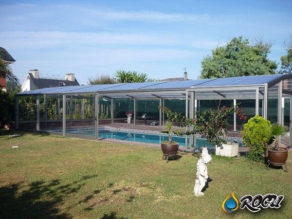 Piscina encerrada en una estructura rectangular con paredes de vidrio y marco de metal, ubicada en un patio trasero con césped bajo un cielo azul.