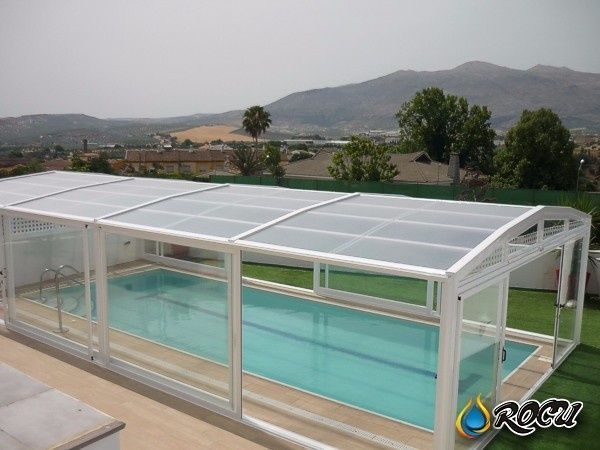 Piscina encerrada en una estructura con techo transparente, ubicada en un patio con un fondo montañoso.
