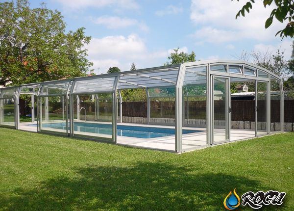 Recinto de piscina con paneles de cristal, marco plateado y techo curvo sobre césped verde.