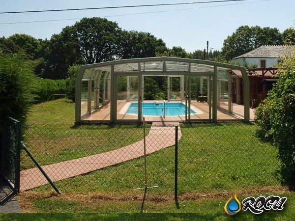 Una piscina rodeada por una estructura transparente se encuentra en un patio cubierto de césped, con un camino que conduce a la entrada.