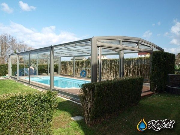 Piscina cerrada con paredes y techo de cristal, ubicada en un patio trasero. Se ven aguas azules, cielo despejado y césped verde.
