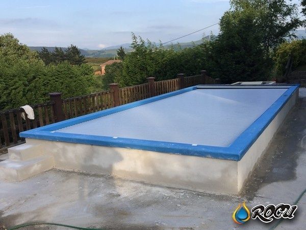 Piscina rectangular con borde azul y cubierta transparente, rodeada de un patio de hormigón y una valla de madera.