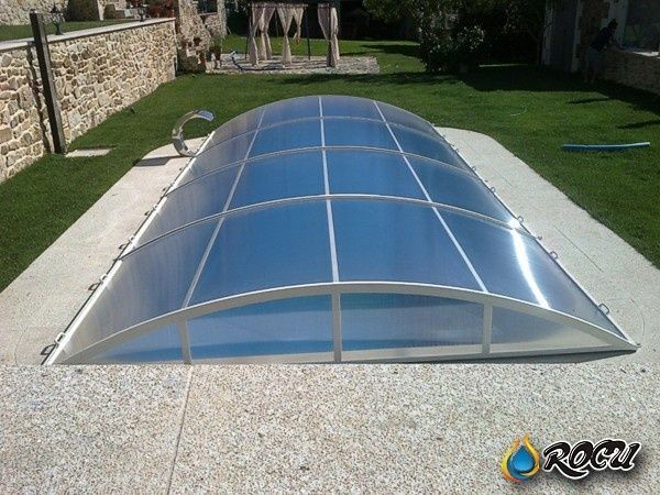 Piscina cerrada con cubierta transparente y arqueada, situada en un patio de piedra con fondo de césped verde.