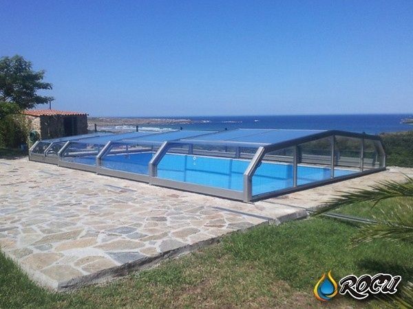 Piscina cubierta por cerramiento retráctil sobre patio de piedra, con vista al mar bajo un cielo azul.