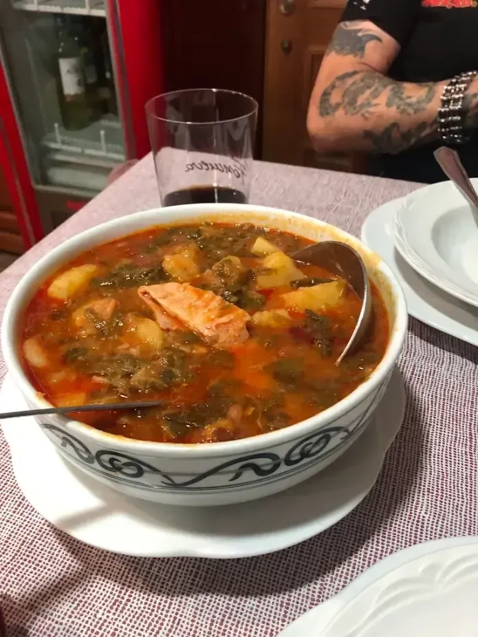 Un tazón de sopa con salmón sobre una mesa junto a un vaso, con un brazo tatuado al fondo.