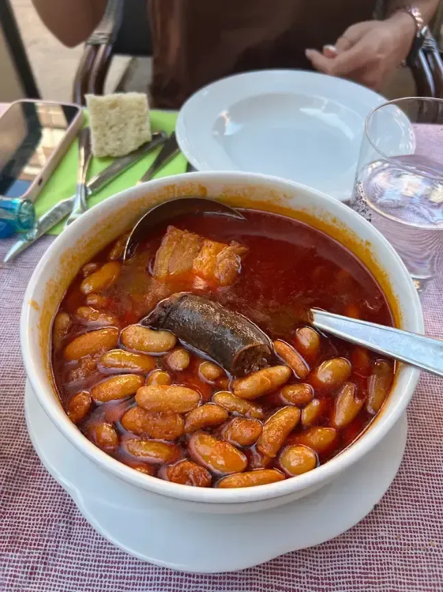 Un tazón blanco de estofado de frijoles rojos con cerdo sobre una mesa, con pan y un vaso cerca.