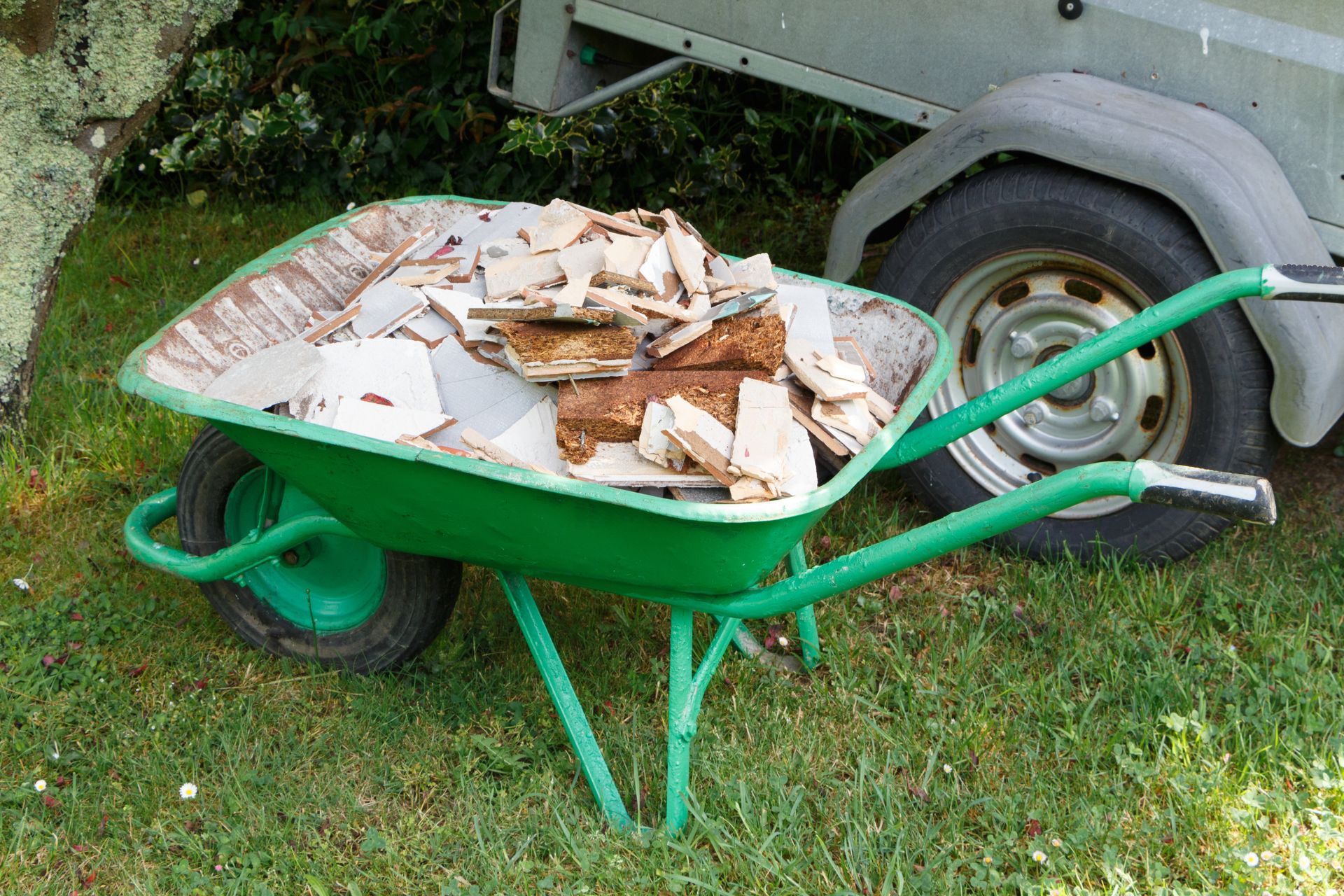 Brouette verte remplie de débris sur l'herbe à côté d'une remorque.