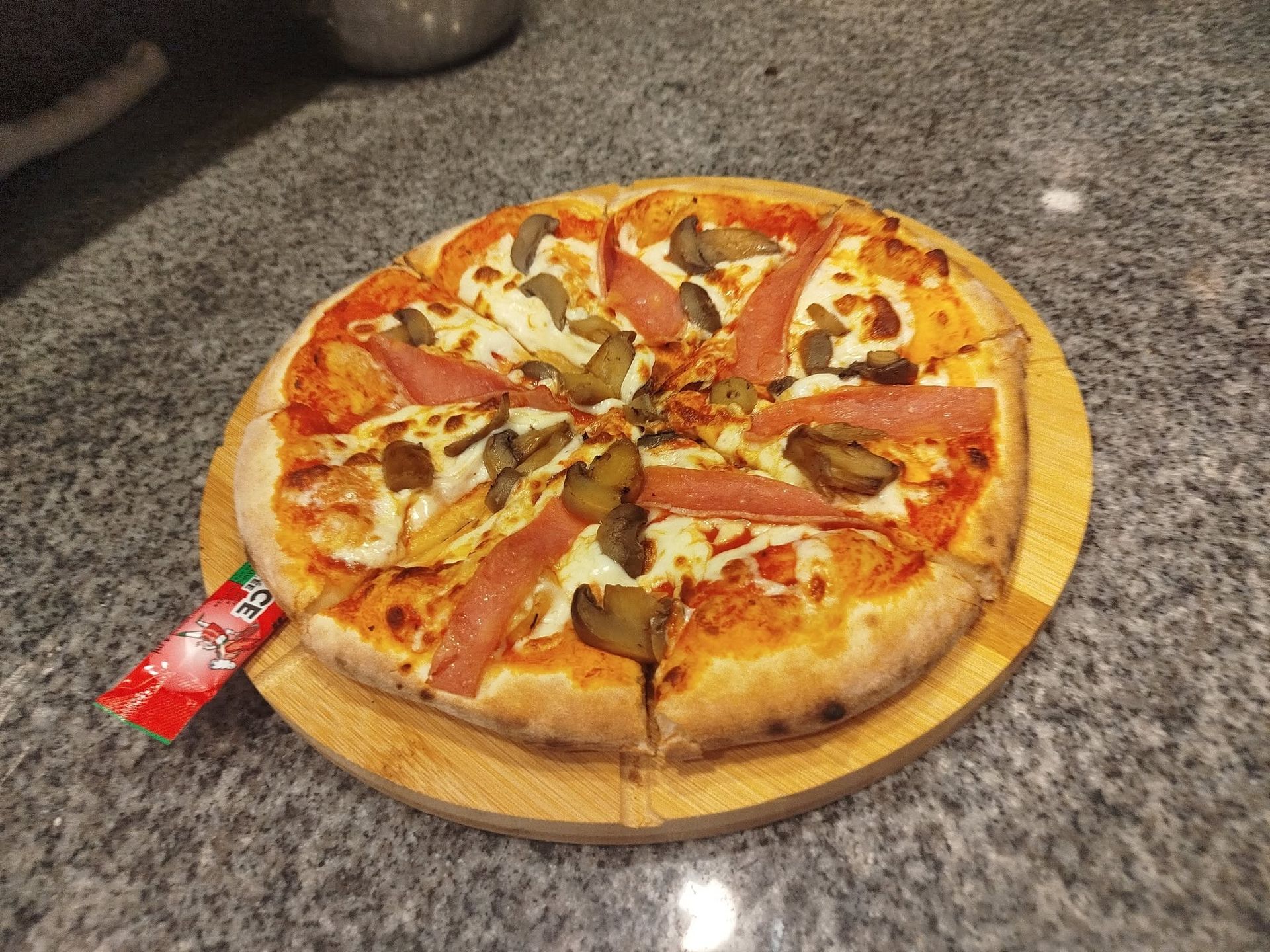 Une pizza sur une planche en bois posée sur le comptoir de la cuisine, avec un emballage rouge à côté.