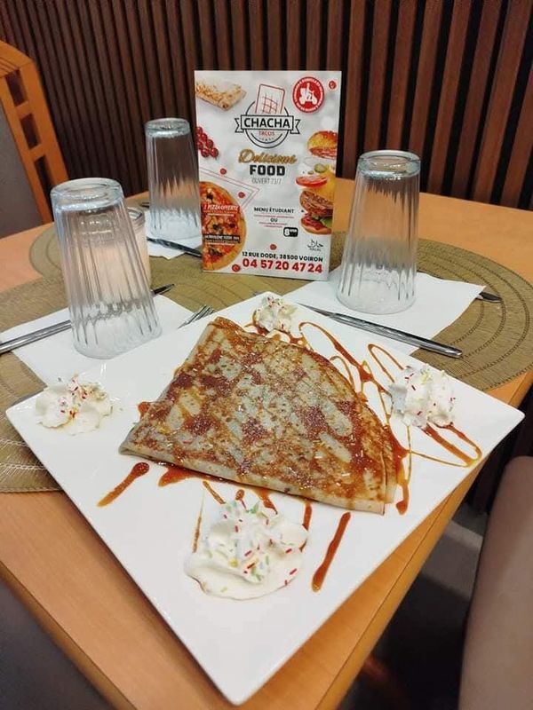Crêpe en dessert, servie sur une assiette blanche avec de la crème fouettée et un filet de caramel, à une table de café avec des verres et un menu.