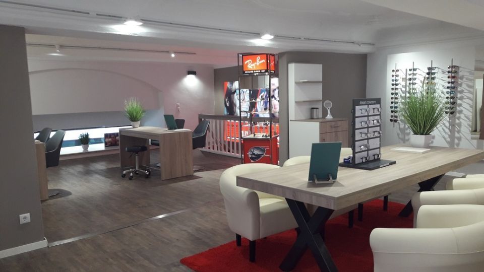 Innenansicht Brillenfachgeschäft: vorne rechts ein Holztisch für vier Personen mit vier weißen Sesseln daran, Brillenaufsteller und Pflanze auf dem Tisch, im Hintergrund Verkaufsbereich