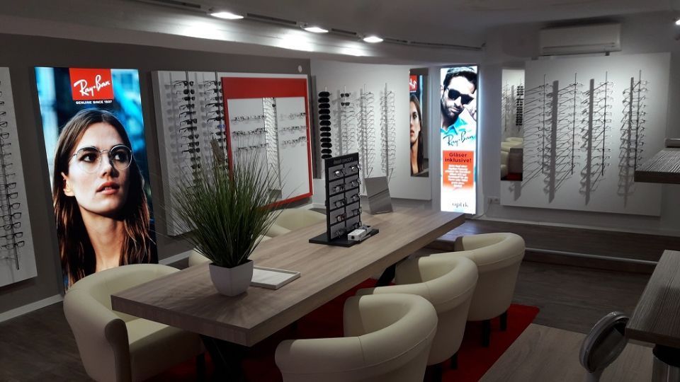 Innenansicht Brillenfachgeschäft: vorne rechts ein Holztisch für vier Personen mit vier weißen Sesseln daran, Brillenaufsteller und Pflanze auf dem Tisch, im Hintergrund Verkaufsbereich