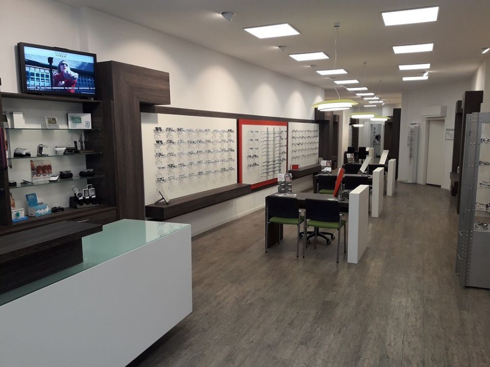 Innenansicht Optiker: links vorn Verkaufstresen, dahinter Brillenwand und Beratungstische, alles in Holzoptik gehalten