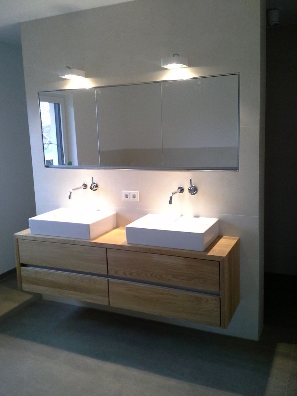 Badezimmeransicht: zwei weiße Waschbecken, darunter Schubladen aus hellem Holz, darüber ein Spiegel