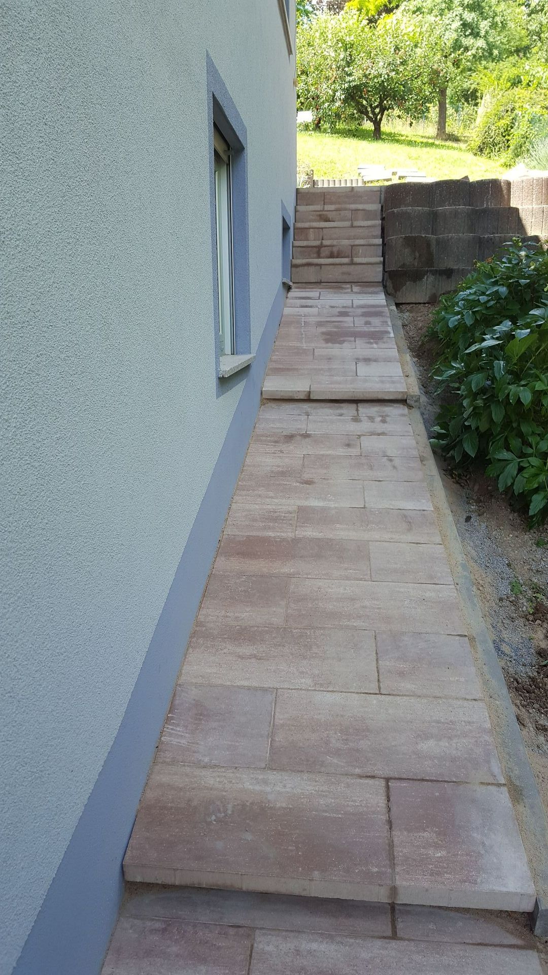 Weg aus Steinpflaster neben einem Gebäude mit Stufen, die nach oben zu einem Terrassengarten führen.
