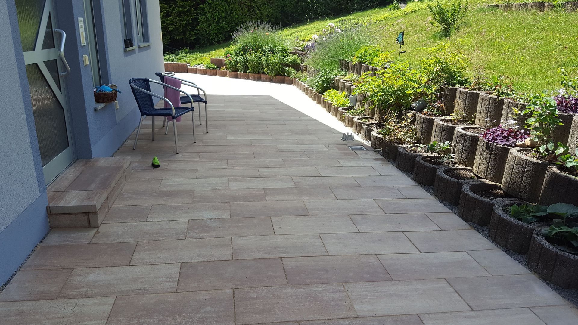 Steinterrasse mit Stühlen, Blumen und Grünpflanzen neben einem Haus.