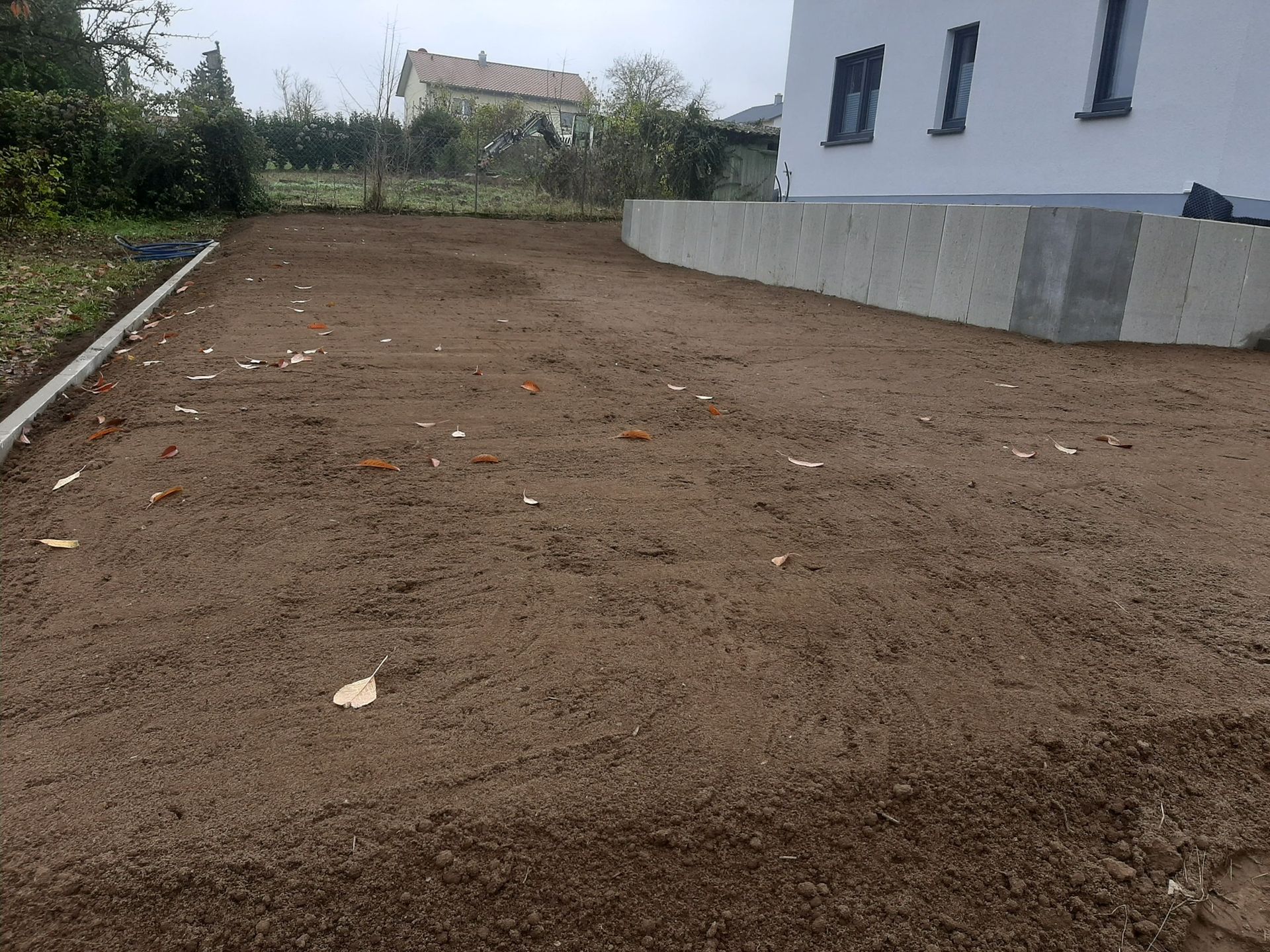 Ein Hof mit frisch gepflügter brauner Erde, neben einem weißen Haus mit Betonstützmauer.