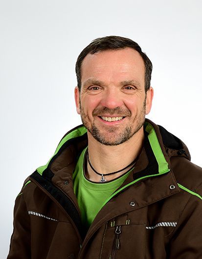 Mann mit kurzen braunen Haaren, lächelnd, trägt eine braune Jacke über einem grünen Hemd.