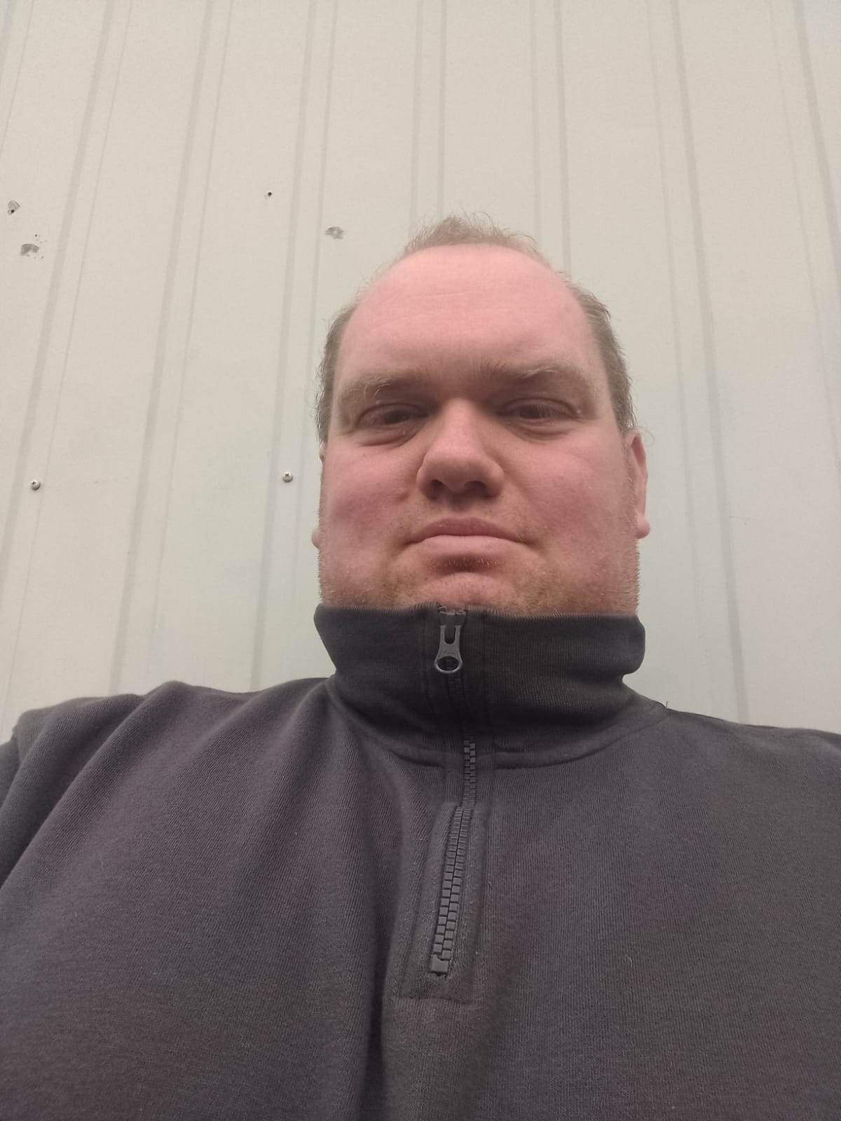 Mann mit heller Haut in einem schwarzen Pullover mit Reißverschluss vor einer schlichten, cremefarbenen Wand.