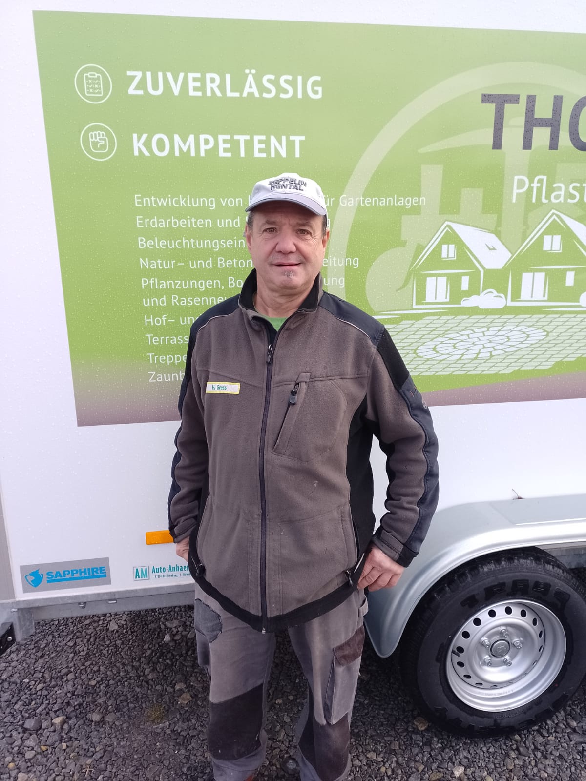 Mann in Arbeitskleidung steht vor einem Anhänger mit grün-weißem Schild.