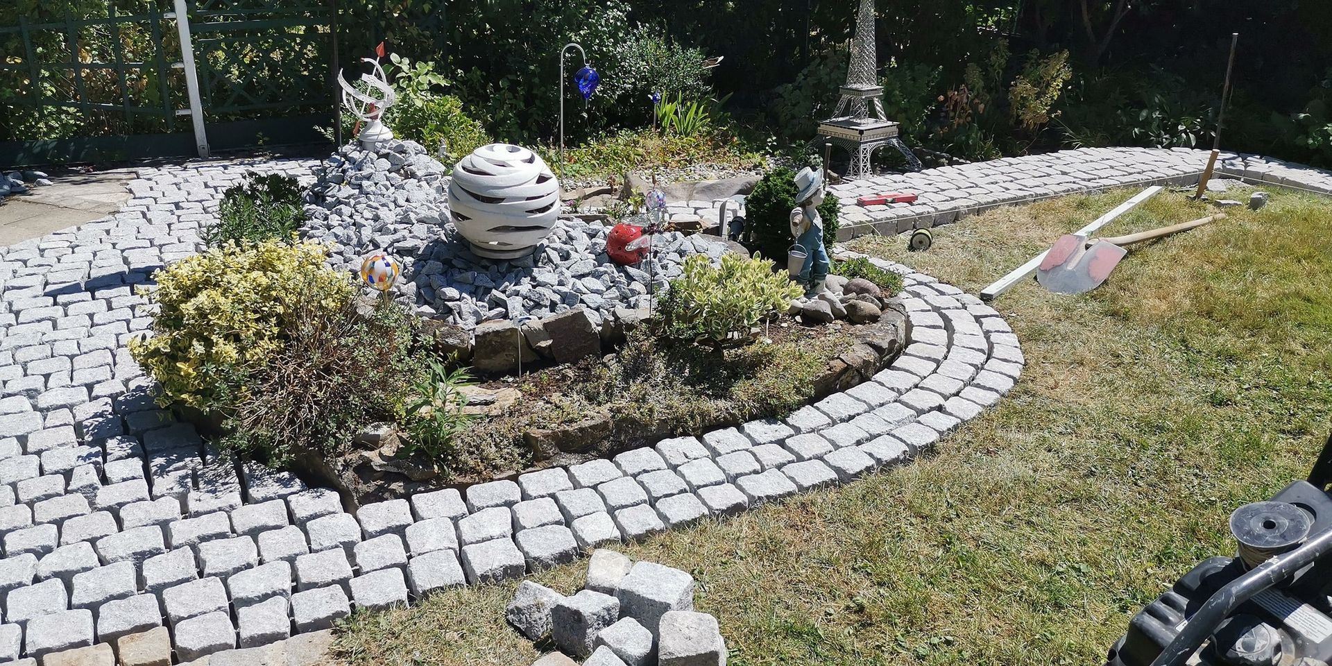 Ein Gartenbeet mit Steinwegen, Pflanzen, Statuen und einer dekorativen Kugel im Freien.