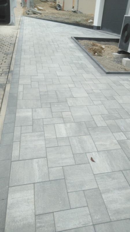 Grau gepflasterter Gehweg, der zu einem Gebäude führt; rechteckiges Steinmuster.