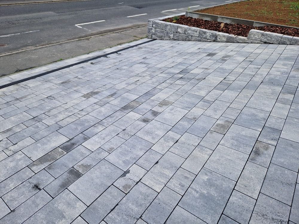 Grau gepflasterte Auffahrt mit kleinen rechteckigen Blöcken, die an eine Straße und ein Blumenbeet aus Stein grenzt.