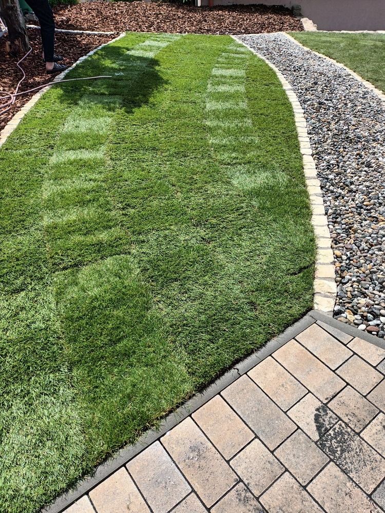 Grüner Rasen, gesäumt von Pflastersteinen, Kiesweg und Mulch, auf dem die Beine einer Person sichtbar sind.