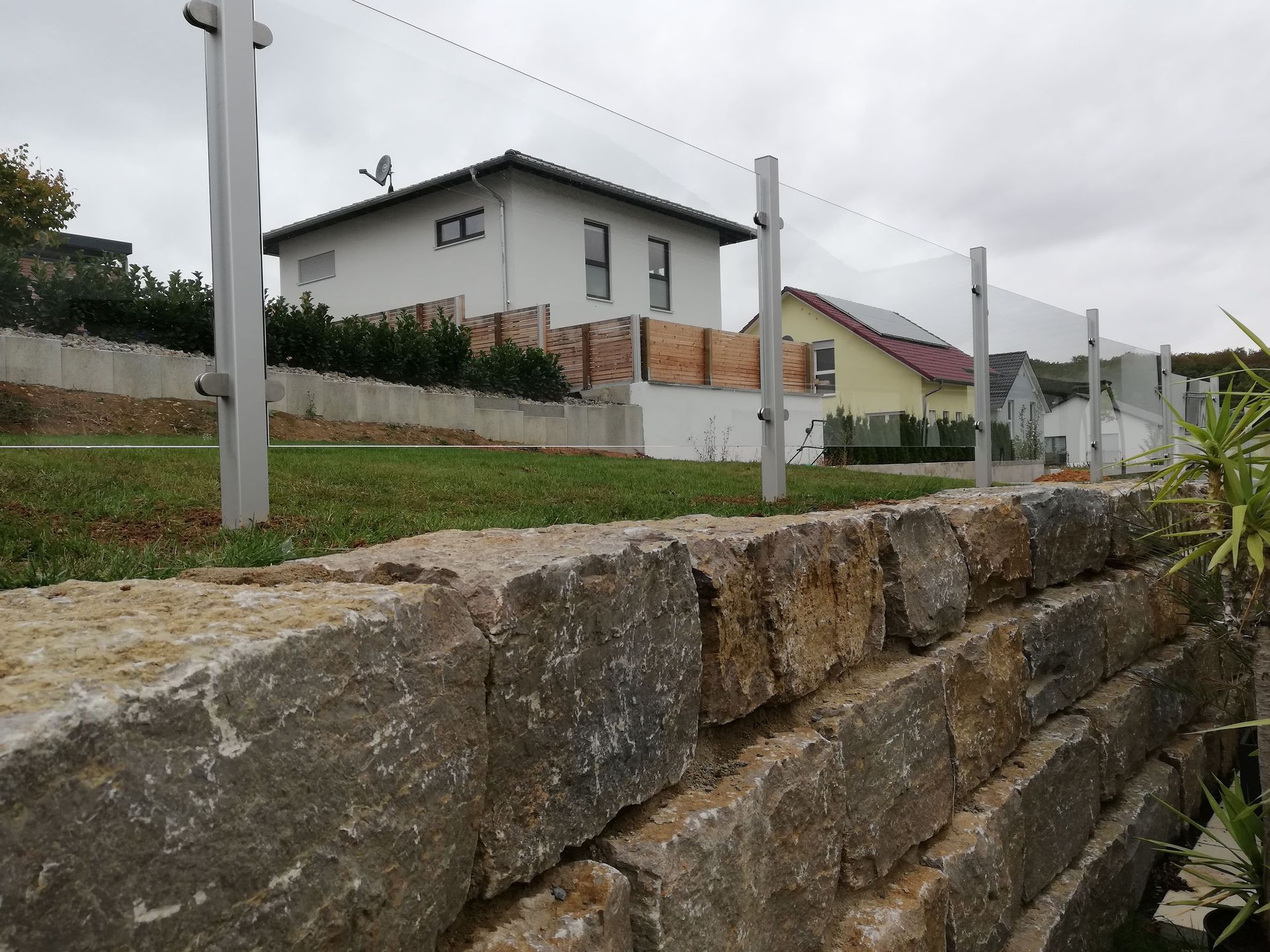 Steinstützmauer mit Metallpfosten und Drahtzaun, Häuser im Hintergrund.