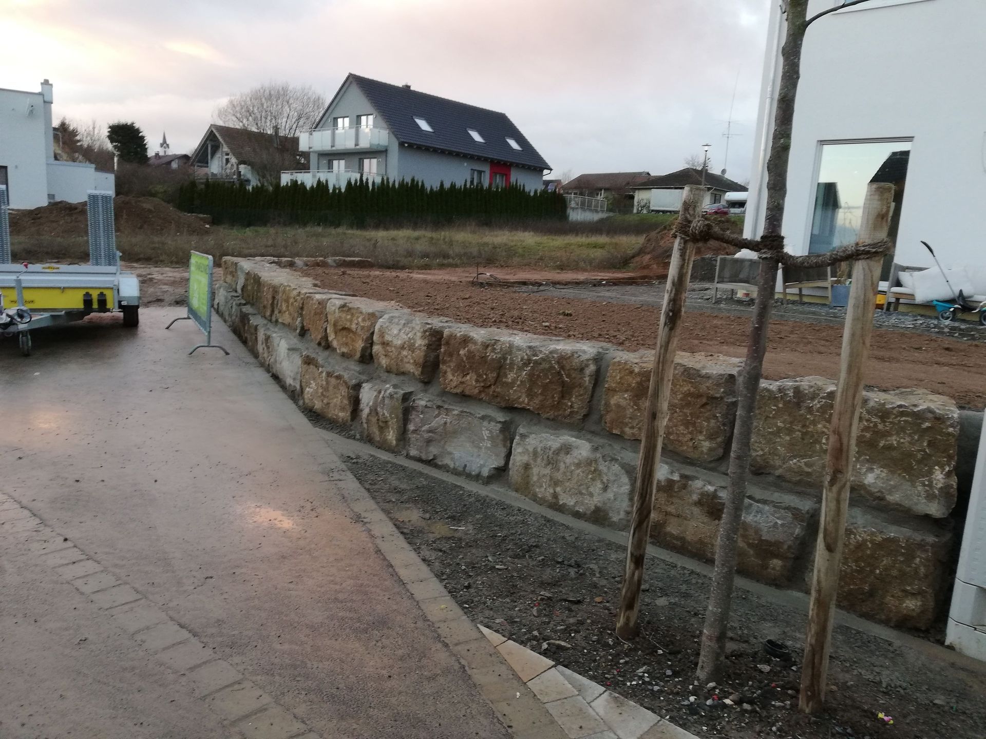 Steinstützmauer entlang einer asphaltierten Straße, im Hintergrund Häuser und Baustelle.