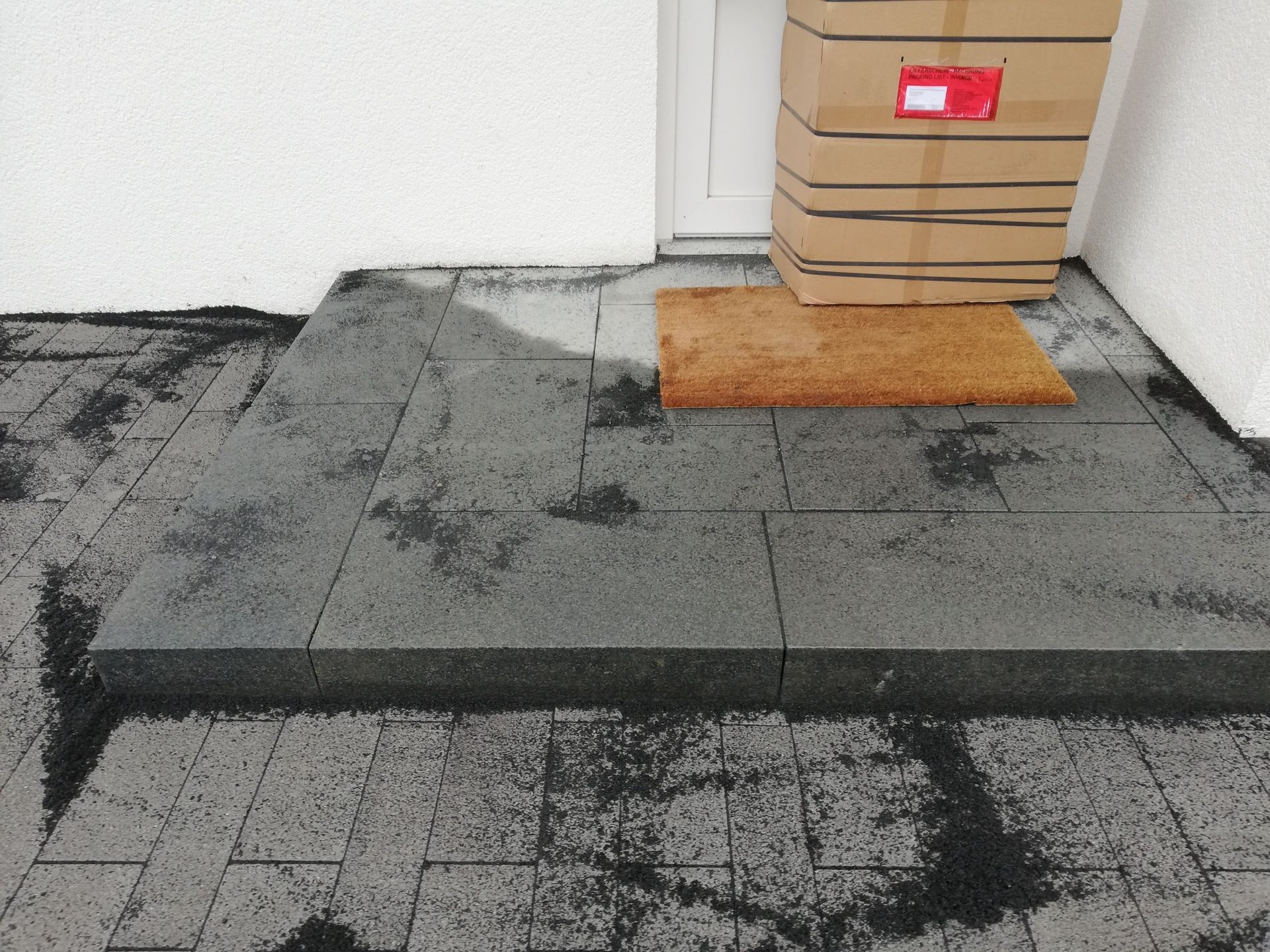 Ein Paket auf einer Fußmatte neben einer Tür auf einem nassen, mit Steinfliesen gefliesten Treppenabsatz.