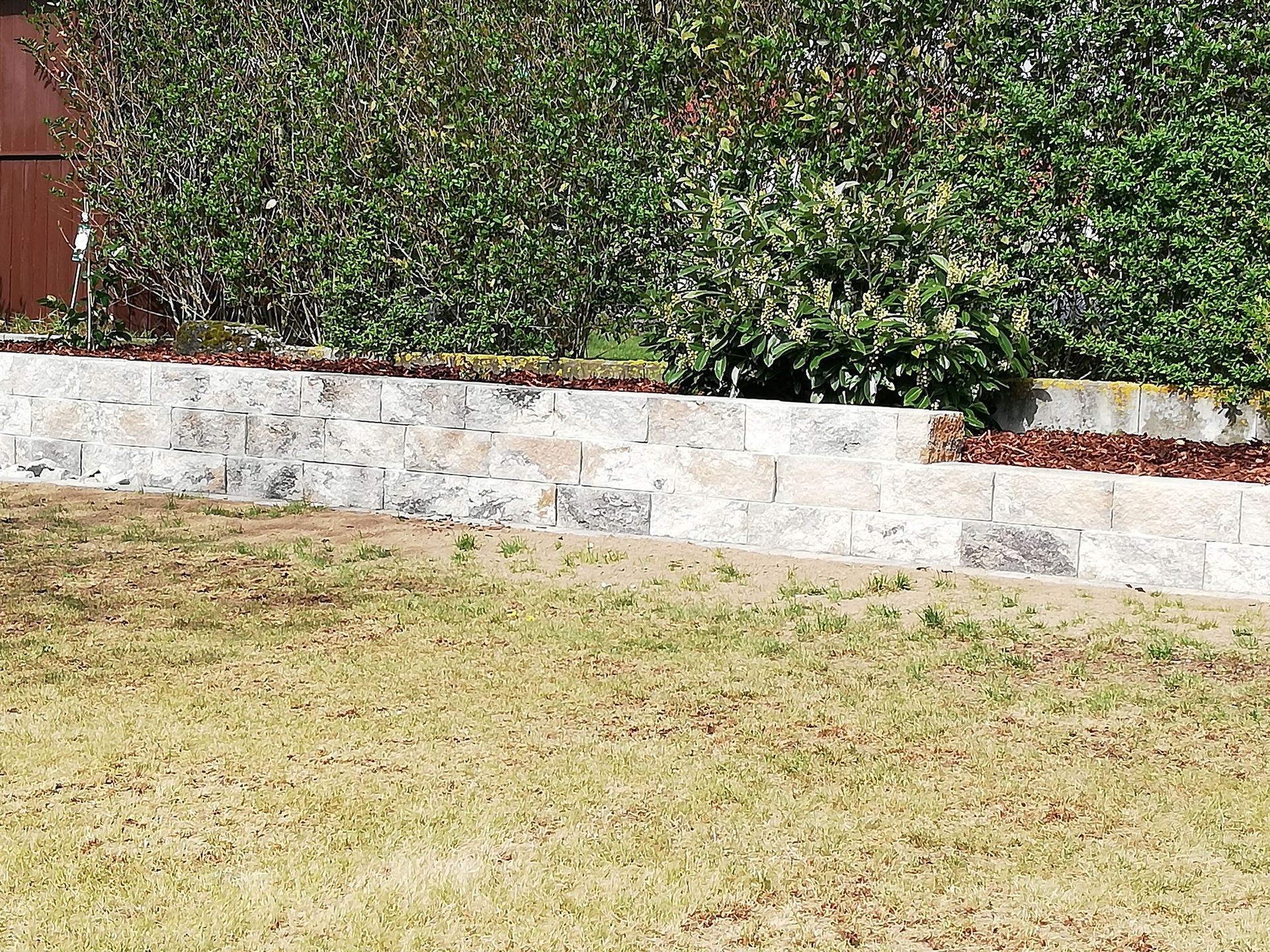Stützmauer aus Stein mit grünen Büschen im Hintergrund und braunem Gras im Vordergrund.